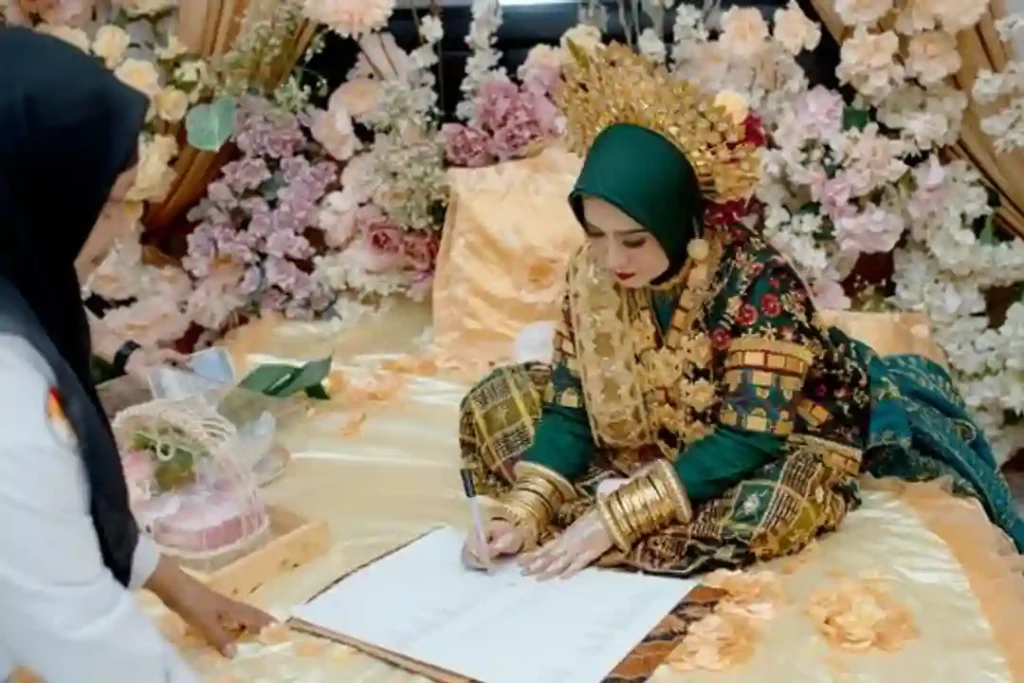 Pengantin wanita mengenakan busana adat Sulawesi Tenggara menandatangani akta nikah di antara dekorasi bunga.