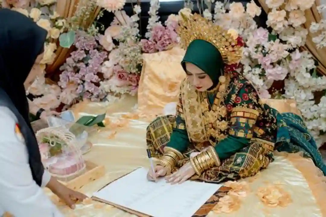 Pengantin wanita mengenakan busana adat Sulawesi Tenggara menandatangani akta nikah di antara dekorasi bunga.